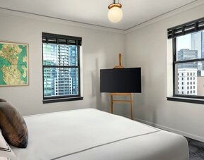 Bedroom with king bed, two windows showing city buildings, and television on stand at Kimpton Palladian Hotel.