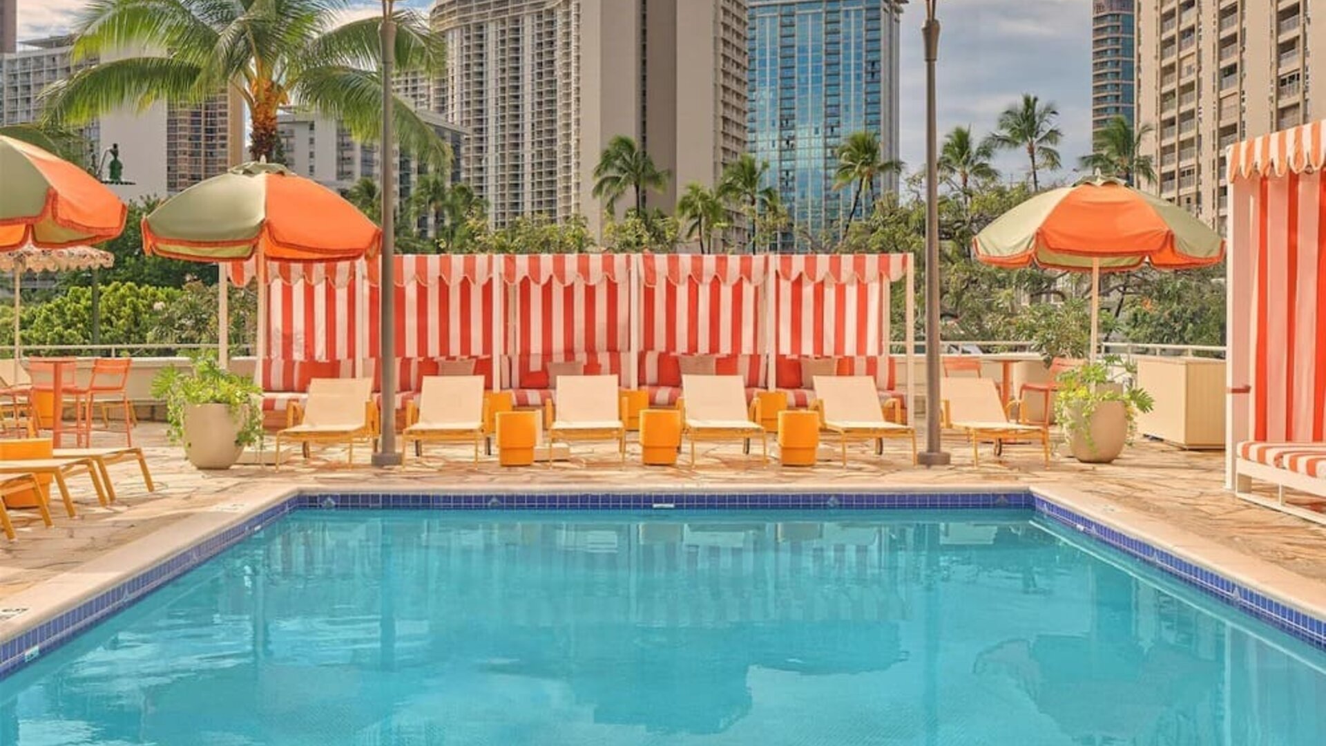 Outdoor pool with lounge chairs, umbrellas, and surrounding city buildings at The Ambassador Hotel Of Waikiki.