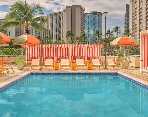 Outdoor pool with lounge chairs, umbrellas, and surrounding city buildings at The Ambassador Hotel Of Waikiki.