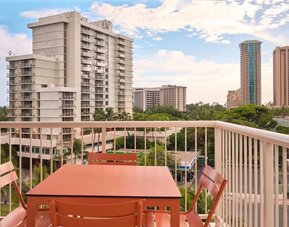 Balcony with table and chairs overlooking city buildings and palm trees at The Ambassador Hotel Of Waikiki.