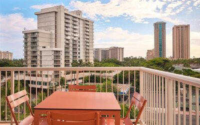 Balcony with table and chairs overlooking city buildings and palm trees at The Ambassador Hotel Of Waikiki.