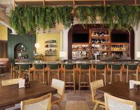 Dining area with bar seating, wooden tables, and hanging greenery decor at The Ambassador Hotel Of Waikiki.
