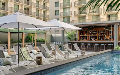 Outdoor pool with sun loungers, umbrellas, tiled deck, and mural wall surrounded by greenery at Romer House Waikiki.