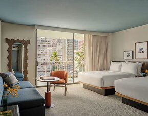 Two double beds with white linens, blue sofa, balcony access, and city view through sliding doors at Romer House Waikiki.