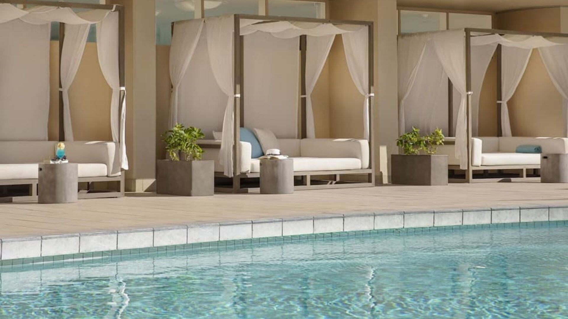 Poolside cabanas with white drapes and cushioned loungers beside outdoor pool at The Twin Fin Hotel.