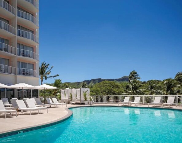 Outdoor pool with lounge chairs, umbrellas, and views of surrounding greenery at Park Shore Waikiki.