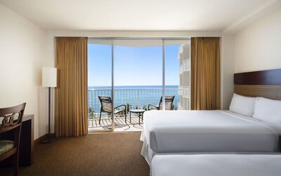Room with two beds, balcony, and ocean view through sliding glass doors at Park Shore Waikiki.