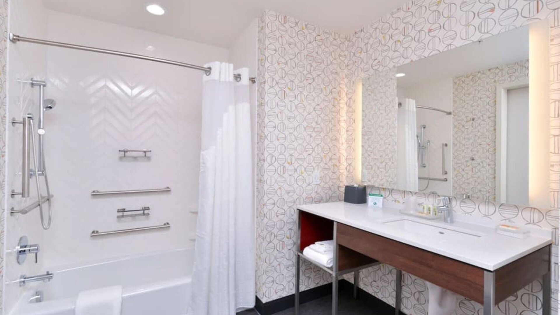 Bathroom with bathtub shower combo, grab bars, patterned wall tiles, and vanity at Holiday Inn & Suites Farmington Hills - Detroit NW By IHG.