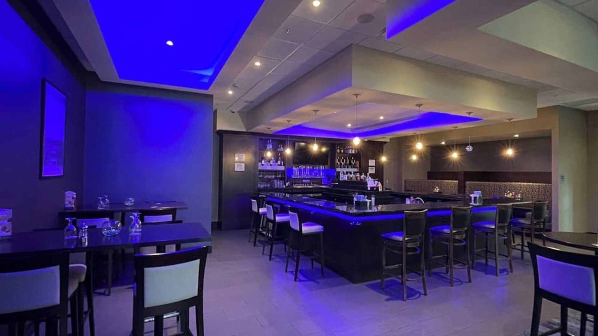 Hotel bar with high stools, ambient blue lighting, and dining tables at Holiday Inn & Suites Farmington Hills - Detroit NW By IHG.