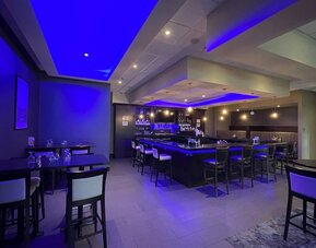 Hotel bar with high stools, ambient blue lighting, and dining tables at Holiday Inn & Suites Farmington Hills - Detroit NW By IHG.