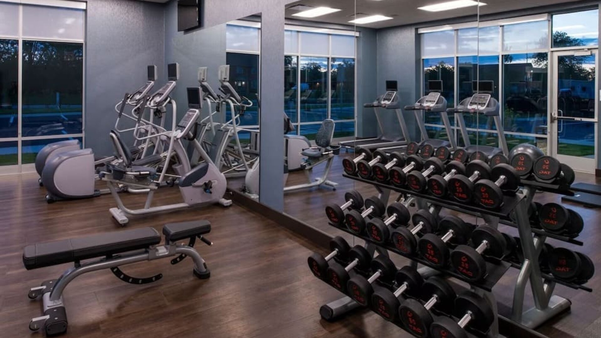 Gym with free weights, cardio machines, mirrored wall, and workout bench at Holiday Inn & Suites Farmington Hills - Detroit NW By IHG.