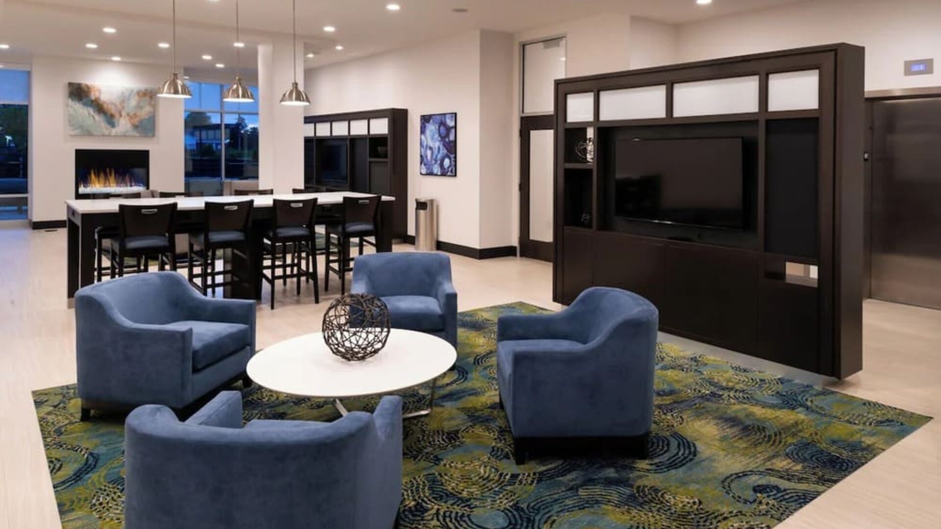 Lounge area with chairs, TV wall, communal table, and fireplace at Holiday Inn & Suites Farmington Hills - Detroit NW By IHG.