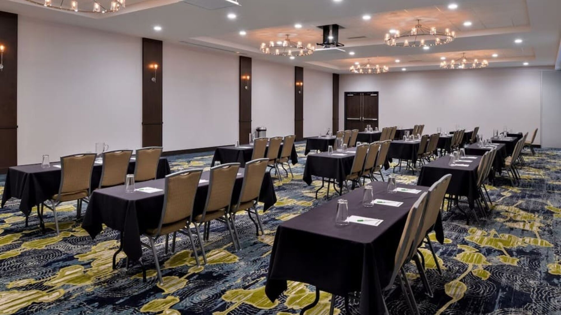 Meeting room with rows of tables, chairs, chandeliers, and patterned carpet at Holiday Inn & Suites Farmington Hills - Detroit NW By IHG.