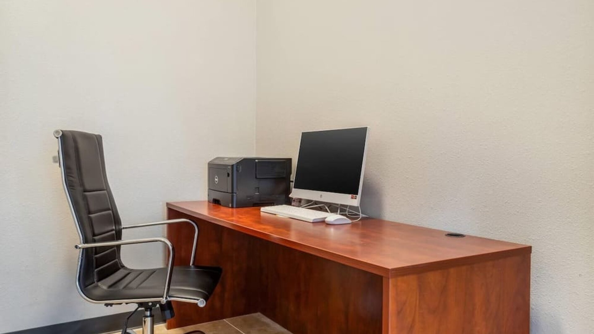 Business center desk with computer, printer, and office chair against a plain wall at Comfort Suites Bush Intercontinental Airport.