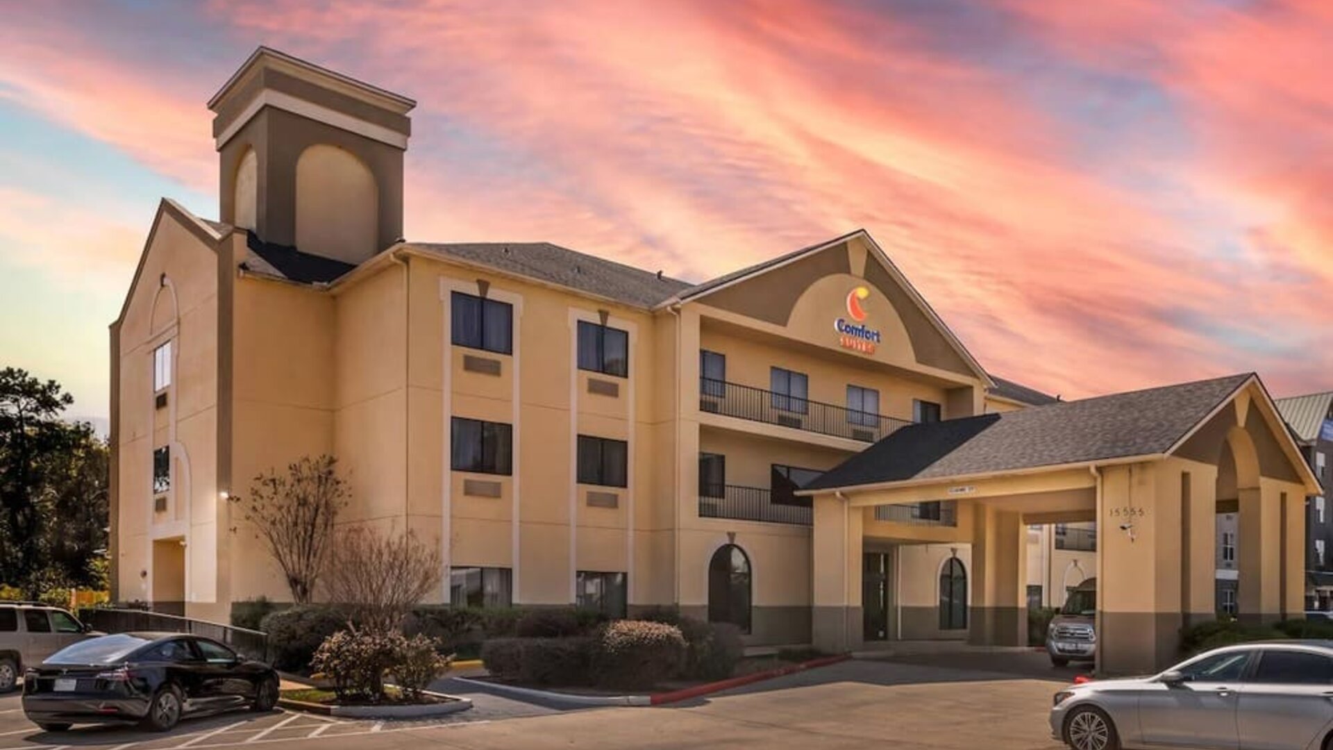 Exterior of hotel building with covered entrance, parked cars, and sunset sky at Comfort Suites Bush Intercontinental Airport.