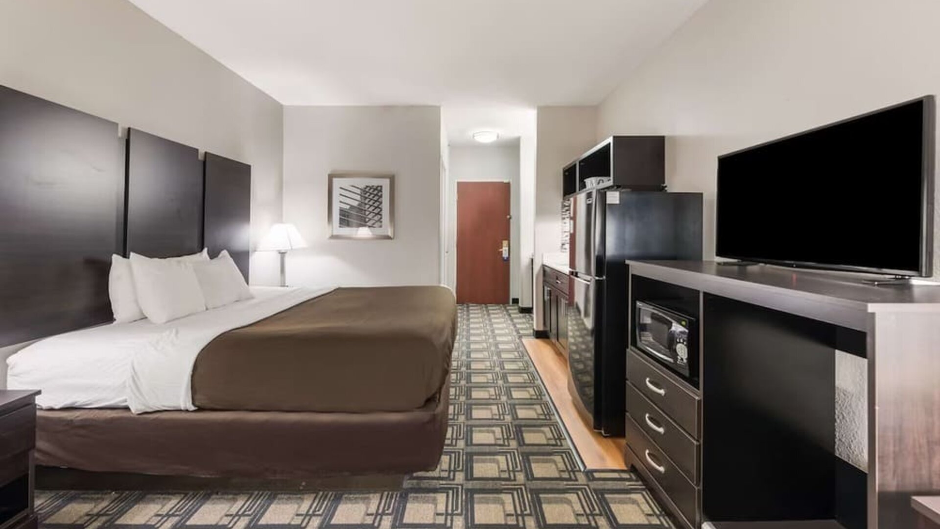 King room with desk area, TV, dresser, and patterned carpet flooring at Suburban Studios Mason Hwy-42.