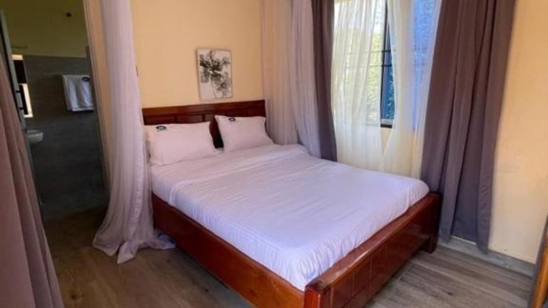 Spacious bedroom with a wooden double bed, canopy frame, and natural light streaming through curtained windows at Sega Lodge.