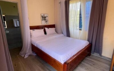 Spacious bedroom with a wooden double bed, canopy frame, and natural light streaming through curtained windows at Sega Lodge.