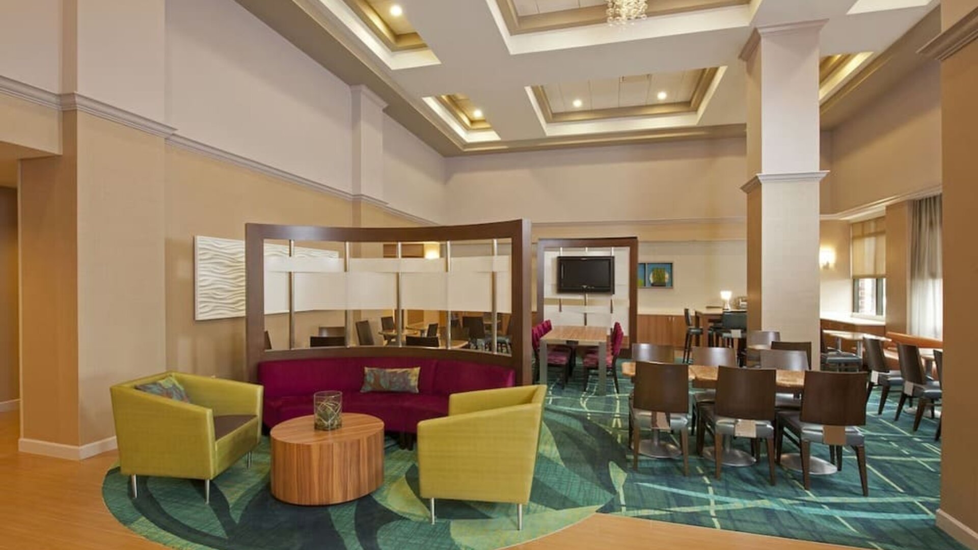 Bright lobby seating area with colorful chairs, round table, and dining tables under coffered ceiling at Marriott SpringHill Suites Naperville/Warrenville.