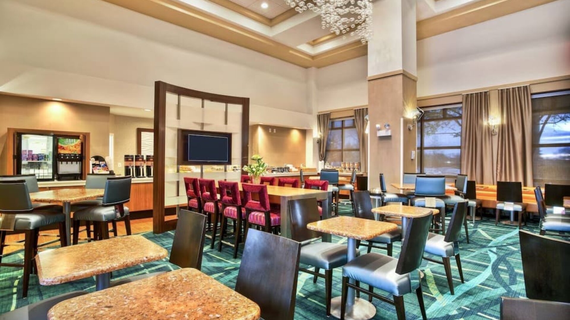 Dining area with multiple tables and chairs, patterned carpet, chandelier lighting, and breakfast counter with beverage station at Marriott SpringHill Suites Naperville/Warrenville.