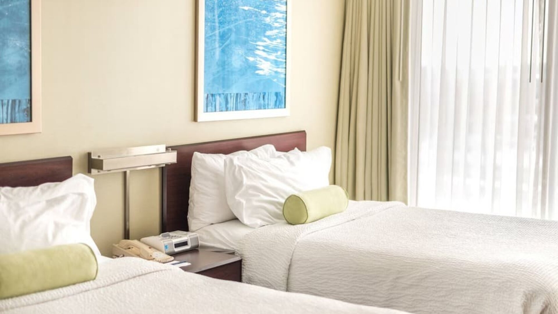 Two double beds with white linens and green bolster pillows, shared nightstand with phone, wall art, and large window with sheer curtains at Marriott SpringHill Suites Naperville/Warrenville.