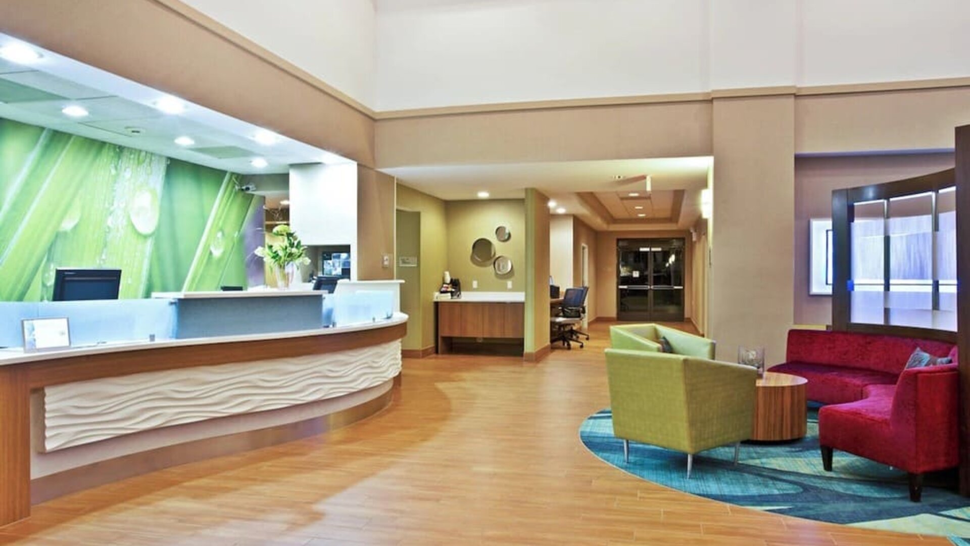 Lobby with curved front desk, green accent wall, wood flooring, and colorful lounge seating area at Marriott SpringHill Suites Naperville/Warrenville.