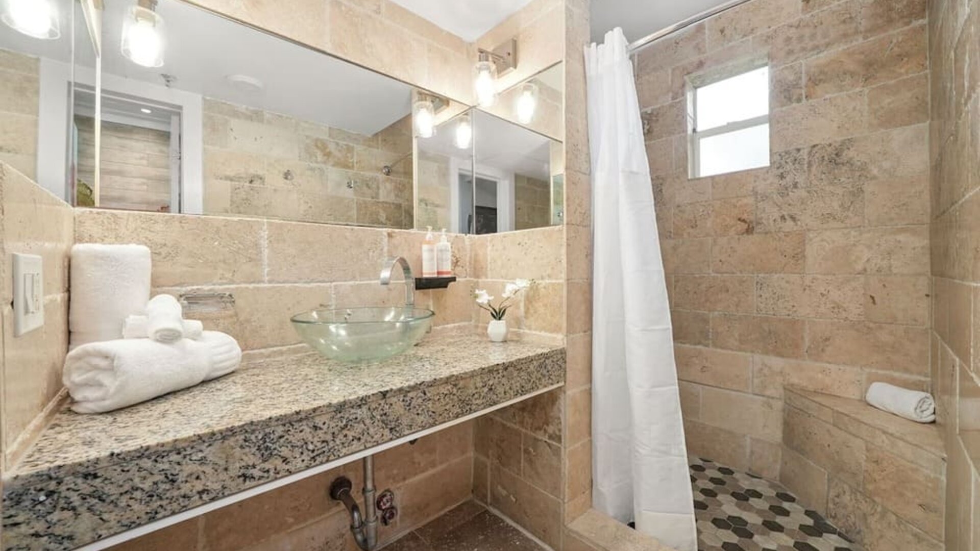 Stone-tiled bathroom with walk-in shower, glass vessel sink on granite counter, and large wall mirror at Chesterfield Hotel & Suites.