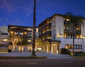 Exterior view at dusk with lit facade, palm trees, and entrance canopy along the street at The Ambrose Hotel Santa Monica.