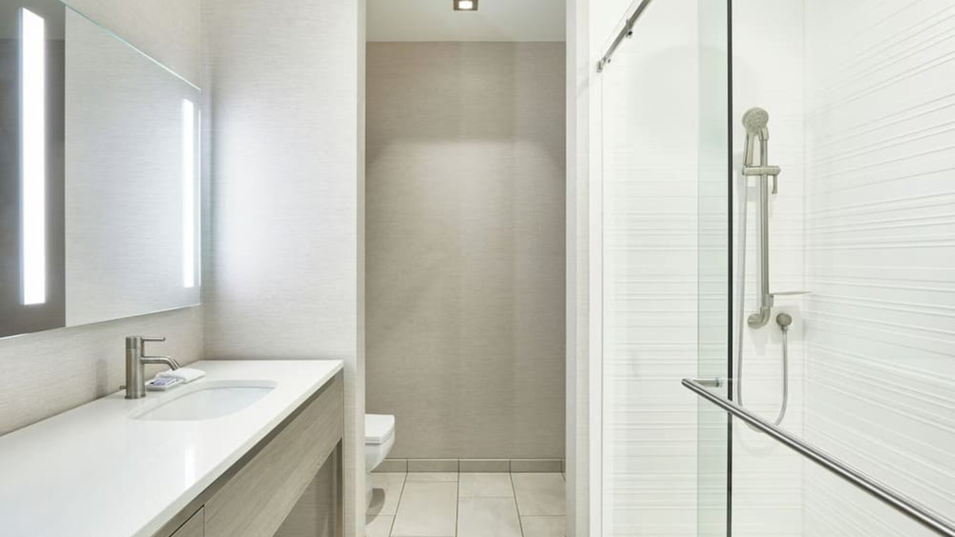 Modern bathroom with long vanity, illuminated mirror, glass shower enclosure, and neutral tile finishes at AC Hotel By Marriott Beverly Hills.