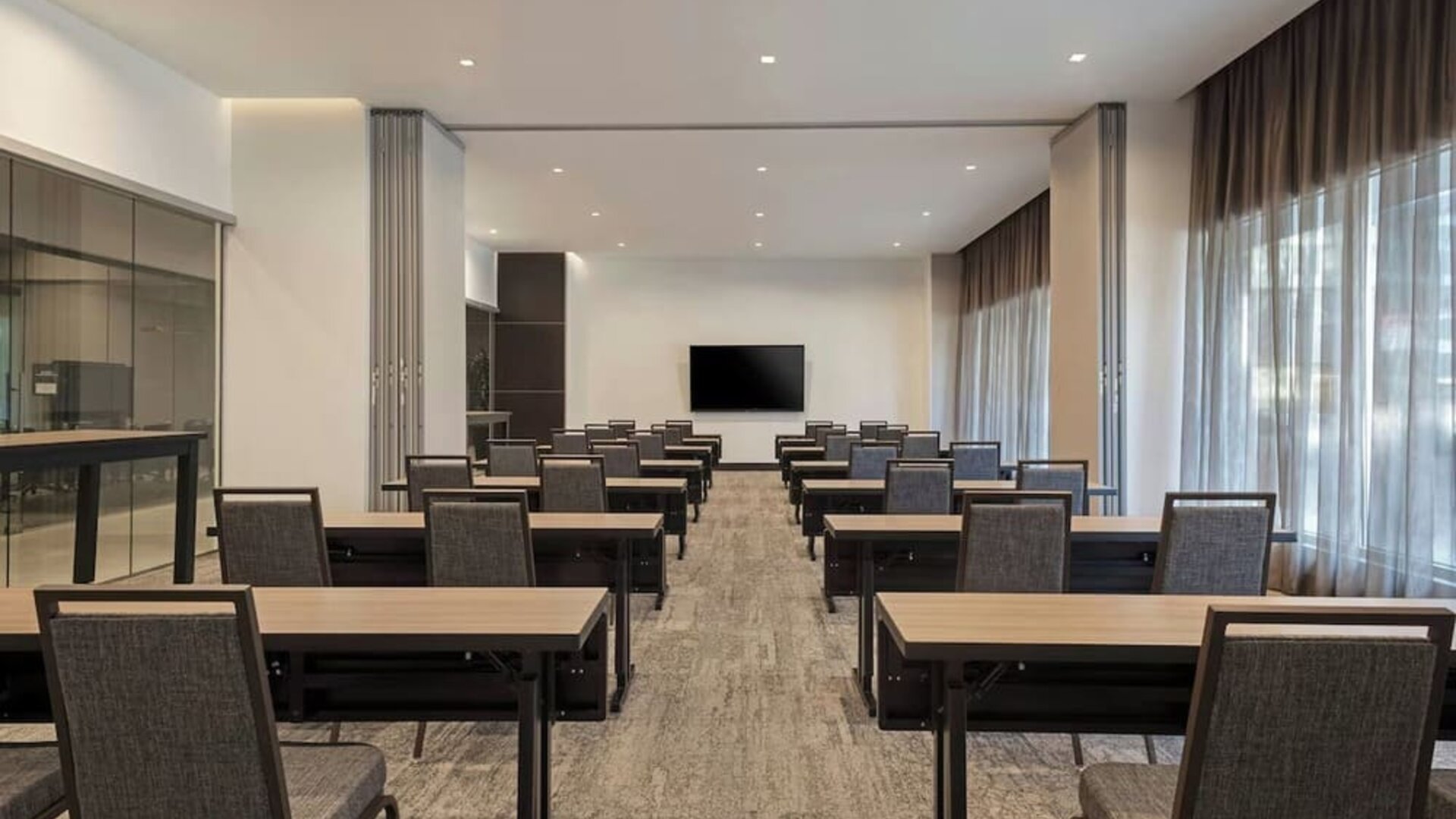 Meeting room with rows of tables and chairs, presentation screen, and large windows with curtains at AC Hotel By Marriott Beverly Hills.