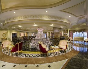 Upper-level lounge with curved railing, chandelier, and clusters of upholstered chairs and tables