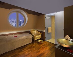 Spa treatment room with massage bed, round window, glass shower, and wooden flooring