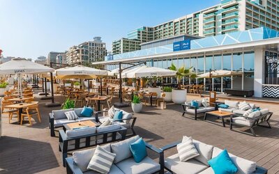 Open-air terrace with cushioned seating, dining tables, umbrellas, and low-rise buildings in background