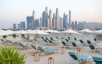 Sandy beach with rows of teal lounge chairs and white umbrellas facing calm water and tall city skyline