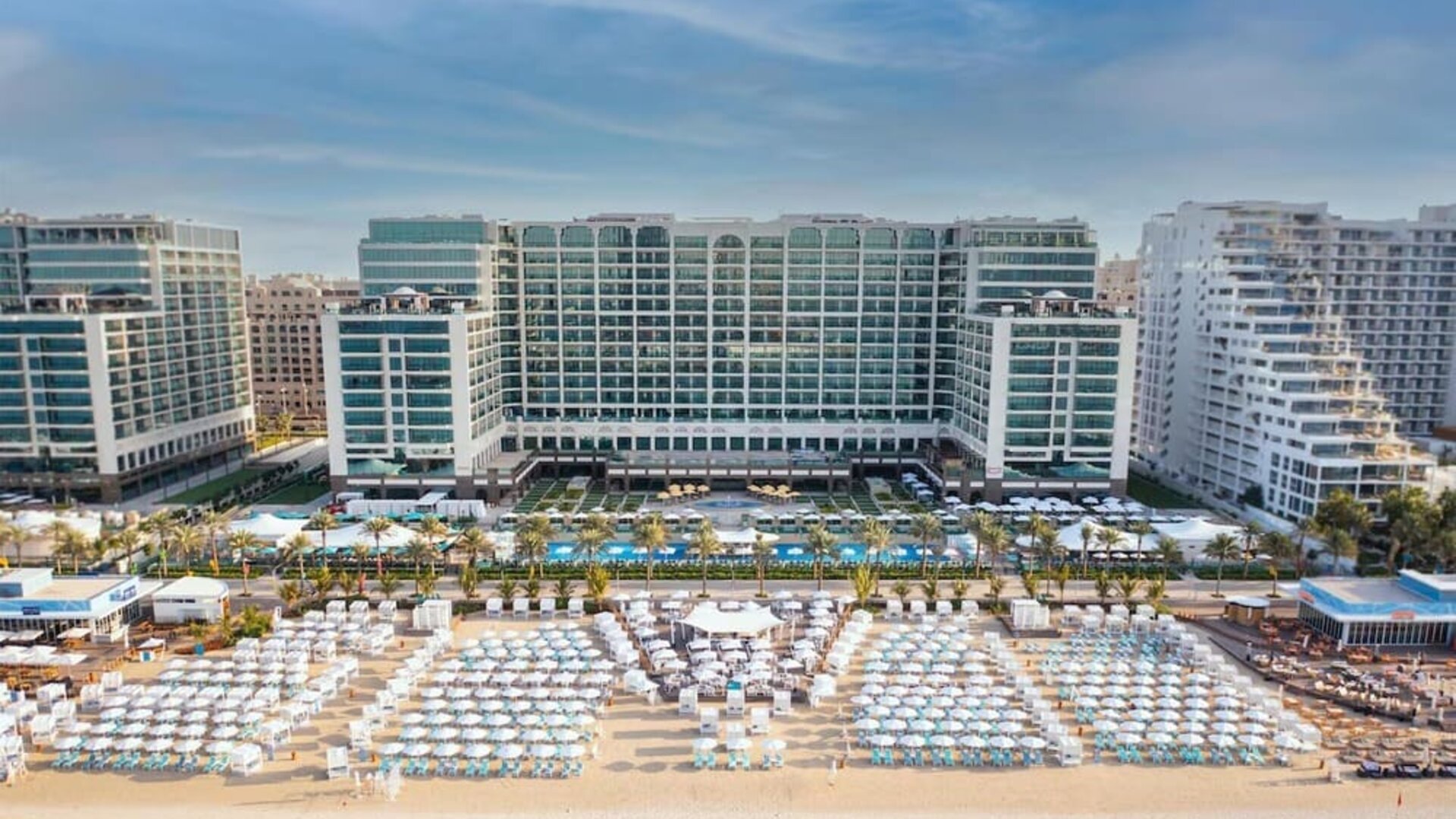 Beachfront hotel exterior with large pool, rows of loungers, and multi-story buildings facing the sea