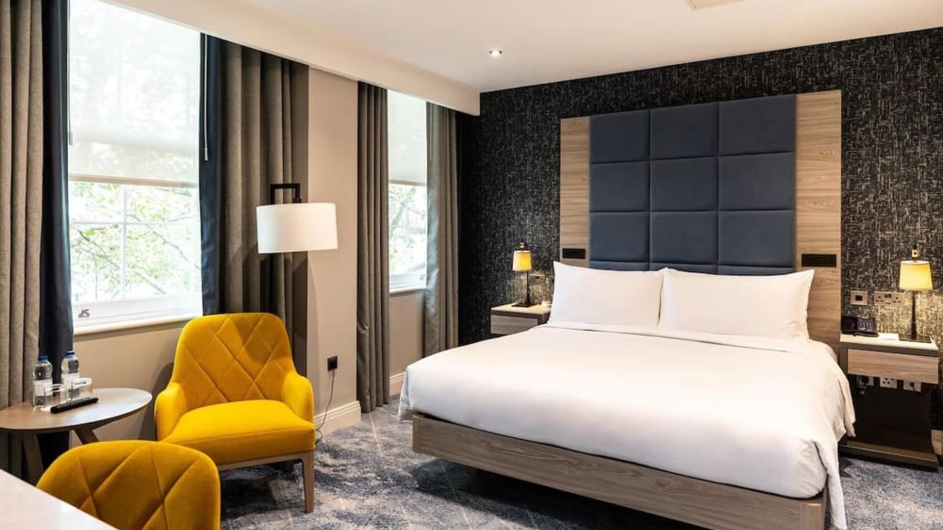 King bed facing two bright yellow armchairs and small round table with water bottles beside a window and desk area
