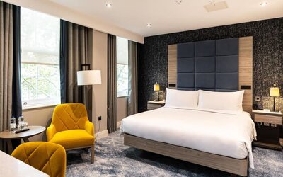 King bed facing two bright yellow armchairs and small round table with water bottles beside a window and desk area