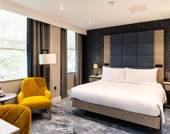 King bed facing two bright yellow armchairs and small round table with water bottles beside a window and desk area