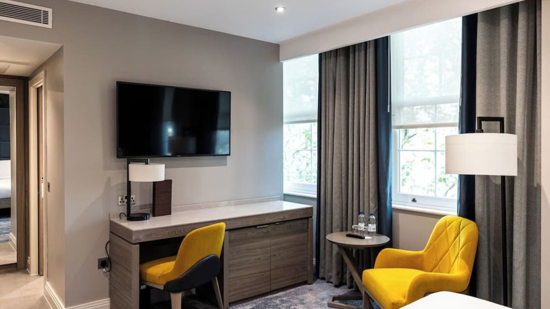 Desk with chair beneath wall-mounted TV, yellow armchair by window, and neutral tones with floor-length curtains