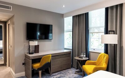Desk with chair beneath wall-mounted TV, yellow armchair by window, and neutral tones with floor-length curtains