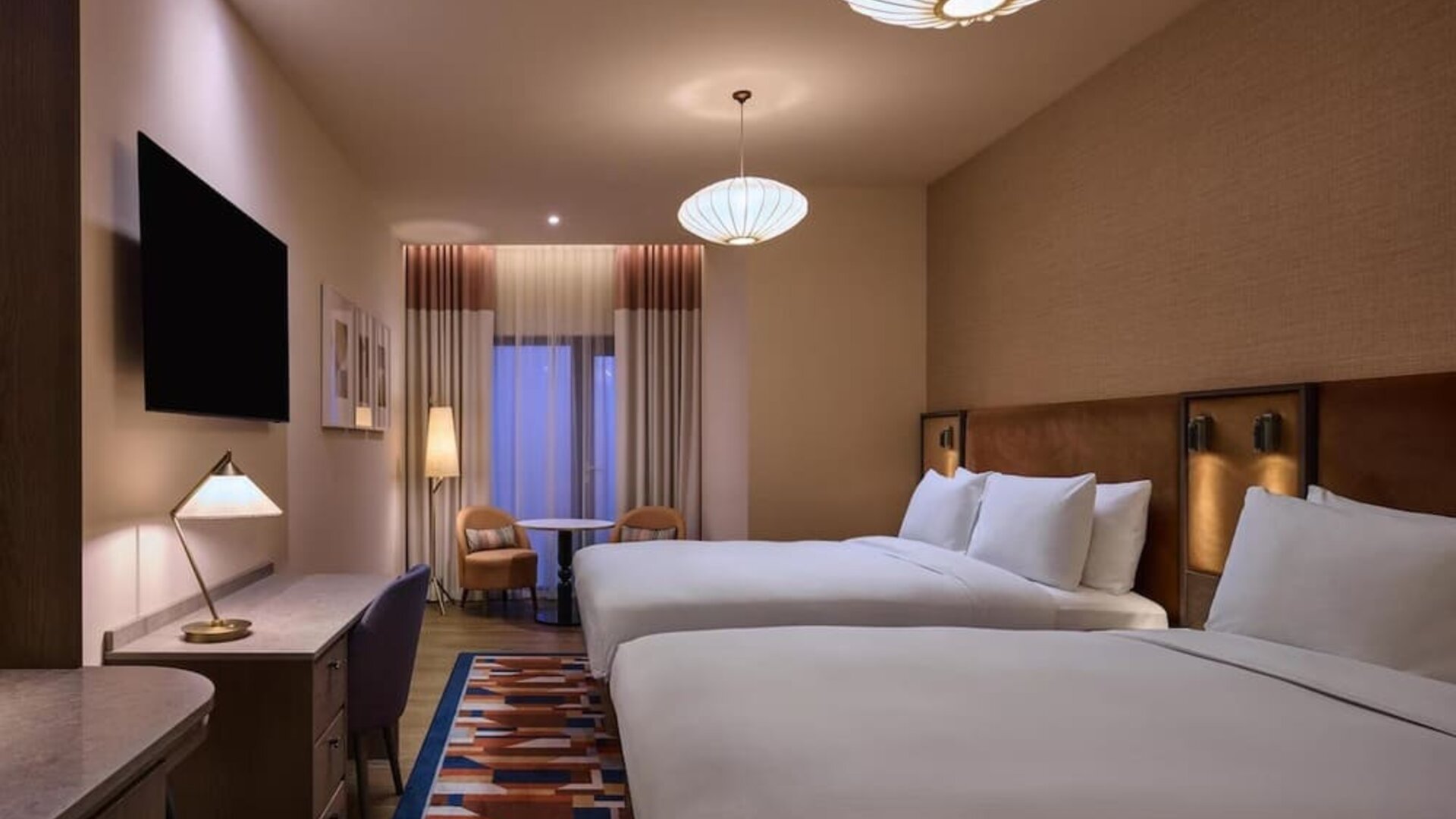Two queen beds with upholstered headboards, patterned rug, desk area, and seating nook near curtained window