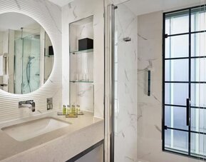 Marble bathroom with glass shower, round mirror above sink, and black-framed frosted window letting in soft light