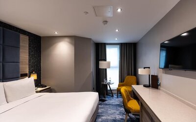 King bed beside long desk and wall-mounted TV, with yellow chair and floor lamp near a narrow window