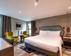 Spacious king room with large bed, padded headboard, desk, and seating area by tall window with dark curtains