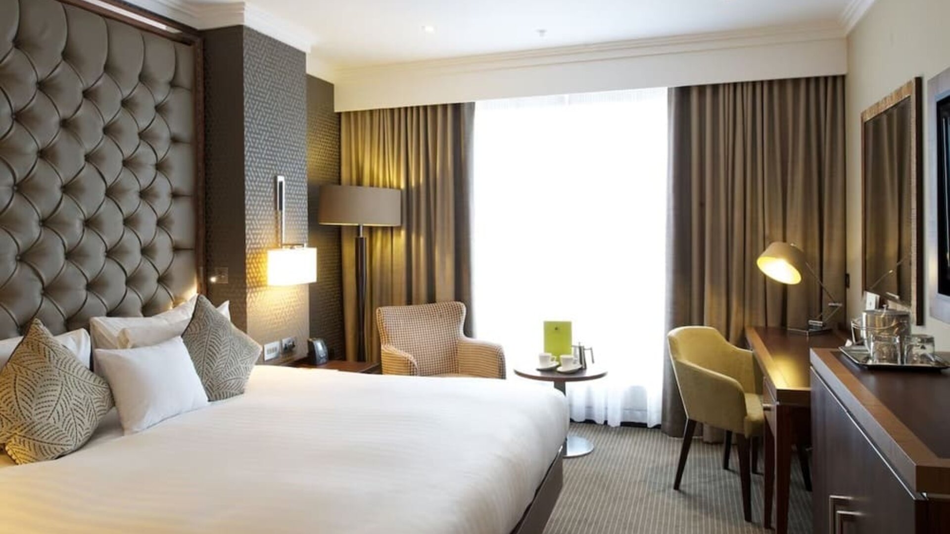 Spacious king room with large bed, padded headboard, desk, and seating area by tall window with dark curtains