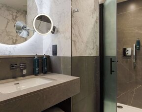 Marble bathroom with illuminated round mirror, modern sink vanity, and glass walk-in shower