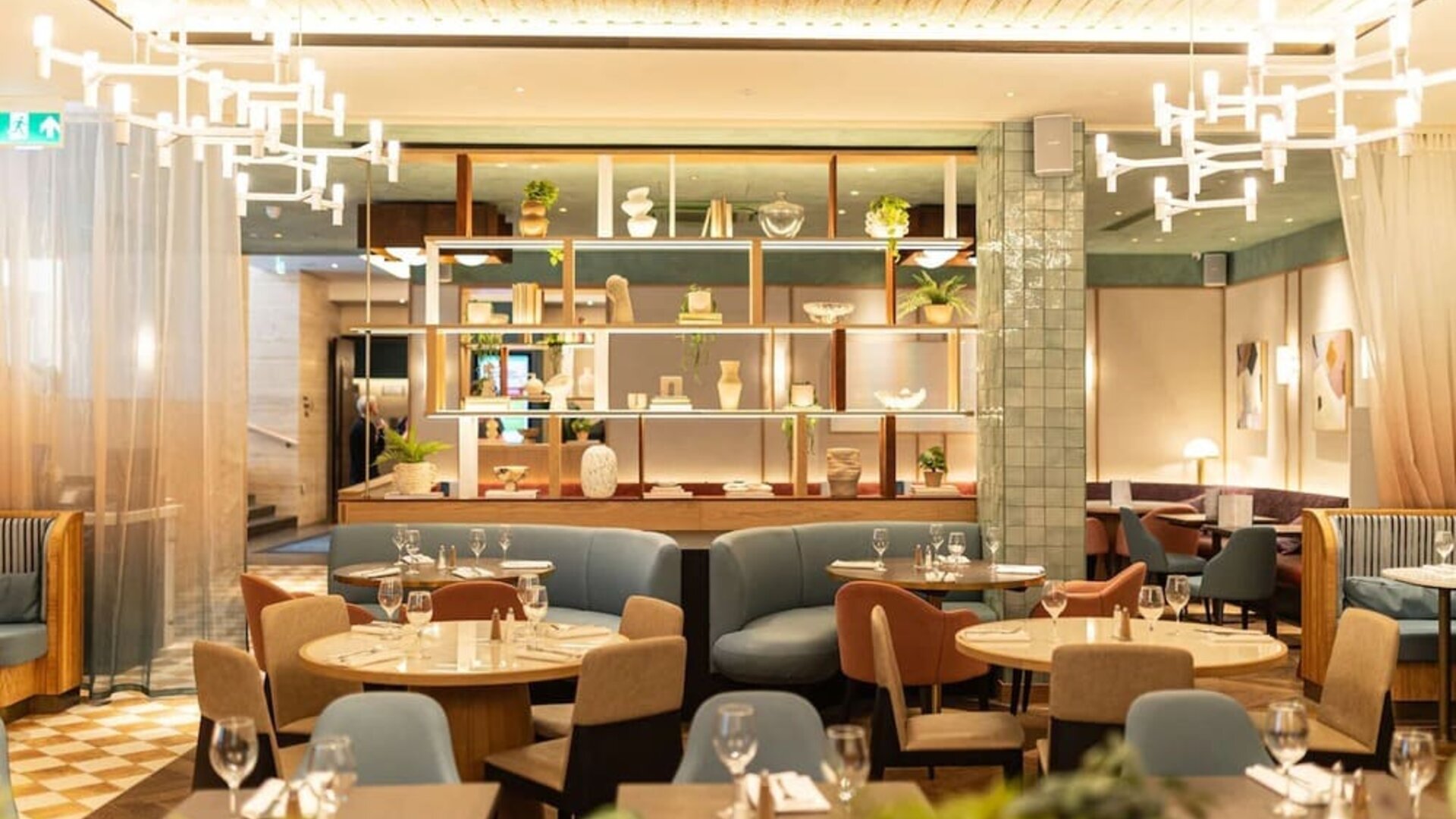 Restaurant dining area with round tables, upholstered seating, decorative shelving, and pendant lights