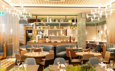 Restaurant dining area with round tables, upholstered seating, decorative shelving, and pendant lights