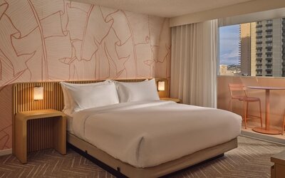 Guest room with king-size bed, modern wood accents, soft ambient lighting, and private balcony seating overlooking Waikiki at The Ambassador Hotel Of Waikiki