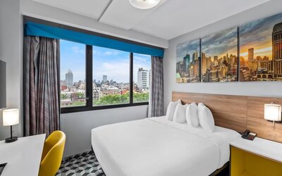 Spacious guest room with large bed, skyline artwork, yellow chair, and expansive city views at Days Inn Chinatown NYC.